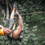 A cool dude looks back at the camera while ziplining above the treetops.