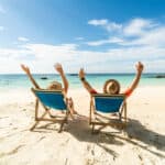 Two people lazing in deck chairs on a beautiful sandy beach through their hands up in the air.