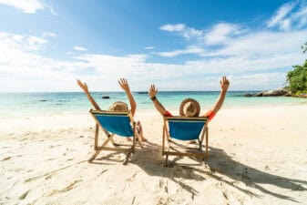 Two people lazing in deck chairs on a beautiful sandy beach through their hands up in the air.