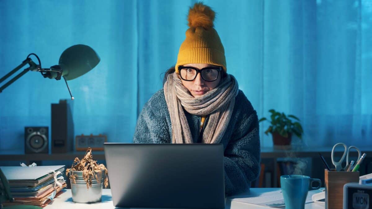 A woman rugged up in winter woollies and a beanie sits frozen at her computer.