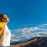 Happy woman stands with raised hands on background of sunset in mountain's range