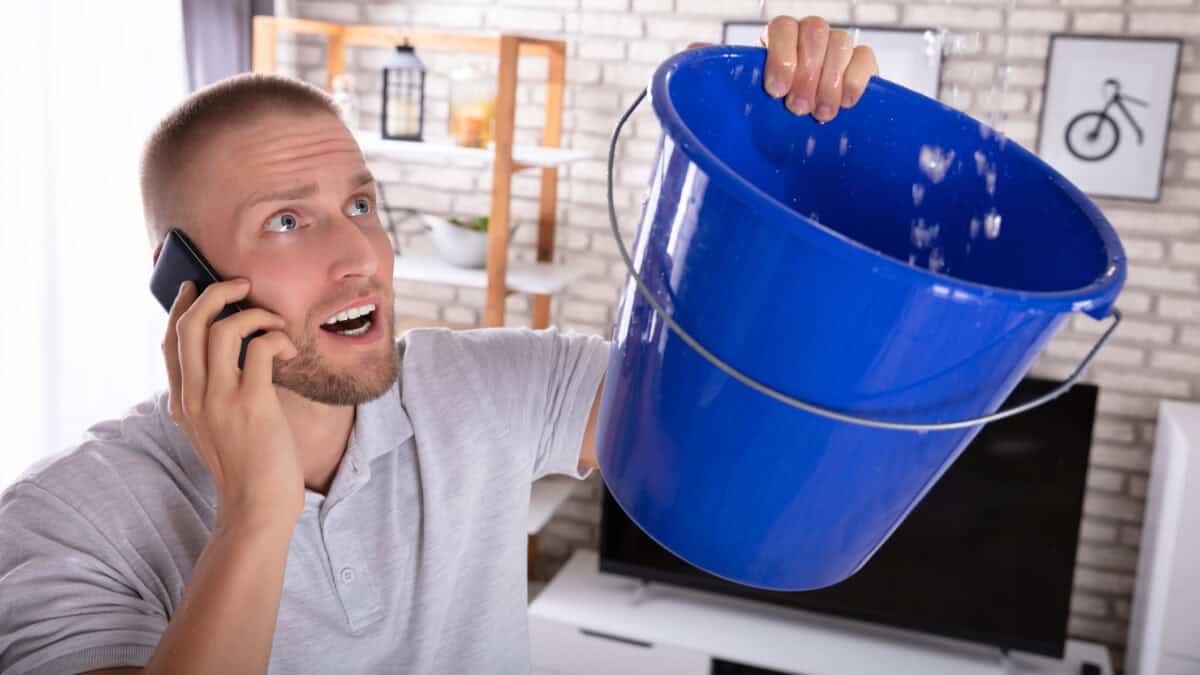 Young man collecting water leakage in bucket while calling plumber on smartphone.