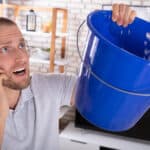 Young man collecting water leakage in bucket while calling plumber on smartphone.