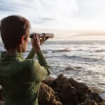 Woman with spyglass looking toward ocean at sunset.