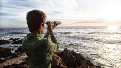 Woman with spyglass looking toward ocean at sunset.