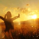 A woman stands in a field and raises her arms to welcome a golden sunset.