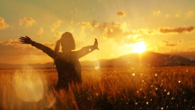 A woman stands in a field and raises her arms to welcome a golden sunset.