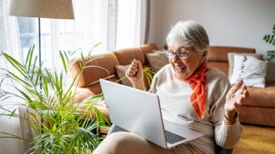 A happy elderly woman smiles and cheers as she looks at good investment news on her laptop.