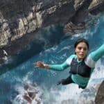 A woman hangs from a cliff with raging waters below.