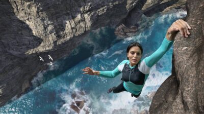 A woman hangs from a cliff with raging waters below.