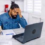 A man holds his head as he looks at his laptop and contemplates more bills to pay.