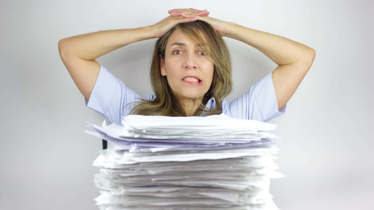 A business woman looks frustrated and angry at a huge stack of paperwork on her desk.