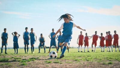 A player kicks a soccer ball to score a goal while players from both teams watch on.