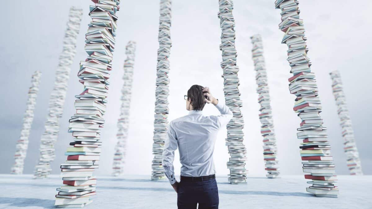 A geeky business man scratches his head as he looks at many stacks of books piled up on the floor.