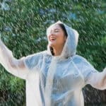 A smiling woman dressed in a raincoat raise her arms as the rain comes down.