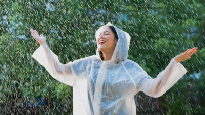 A smiling woman dressed in a raincoat raise her arms as the rain comes down.