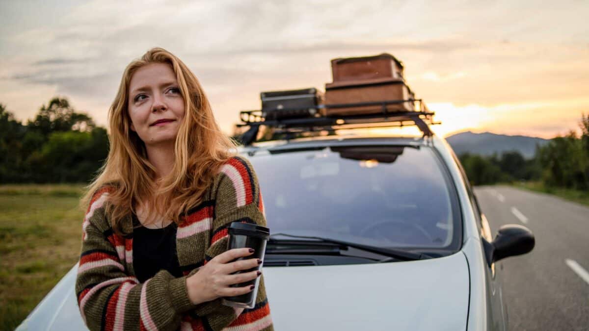 A woman smiles as she stands next to a car loaded with a stack of suitcases on the roof.