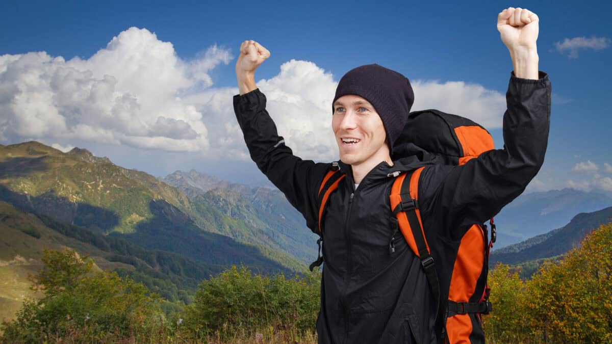 Hiker man backpacker with hands up in the summer mountains with cloudy sky.