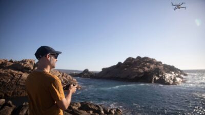 A man wearing a cap flies his drone at the beach.