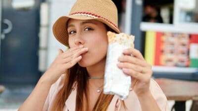 I young woman takes a bite out of a burrito n the street outside a Mexican fast-food establishment.