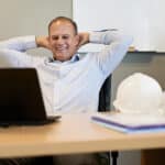 Cheerful businessman with a mining hat on the table sitting back with his arms behind his head while looking at his laptop's screen.