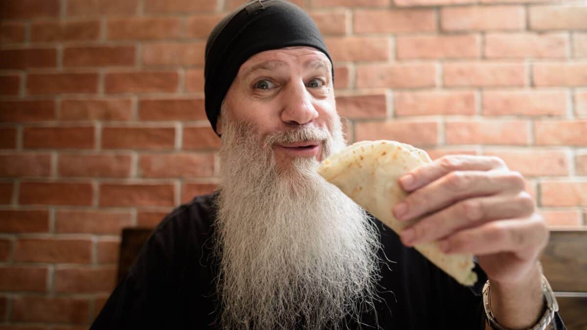 An old dude with a long flowing beard smiles as he bites into a Mexican burrito.