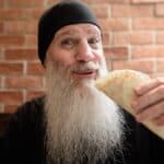 An old dude with a long flowing beard smiles as he bites into a Mexican burrito.