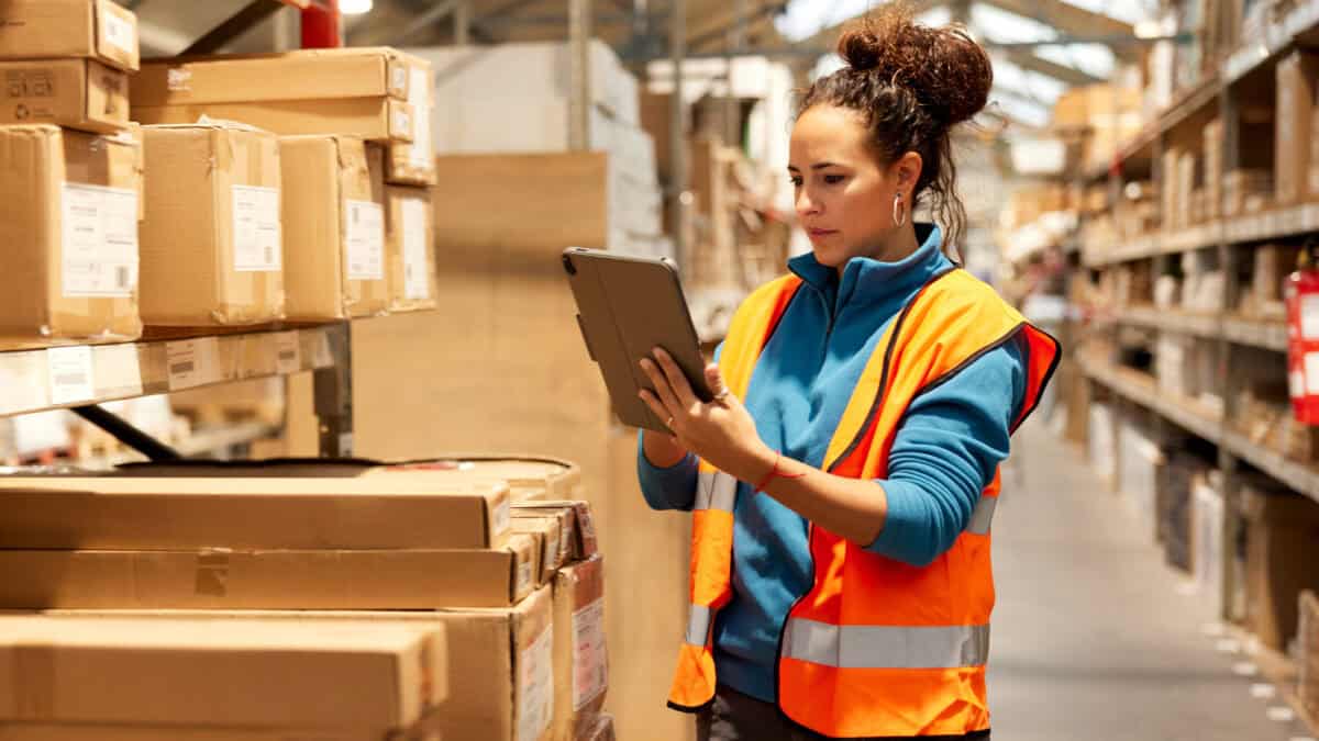 A warehouse worker is standing next to a shelf and using a digital tablet.