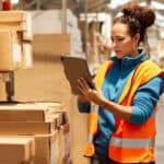 A warehouse worker is standing next to a shelf and using a digital tablet.