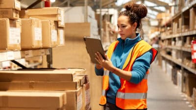 A warehouse worker is standing next to a shelf and using a digital tablet.