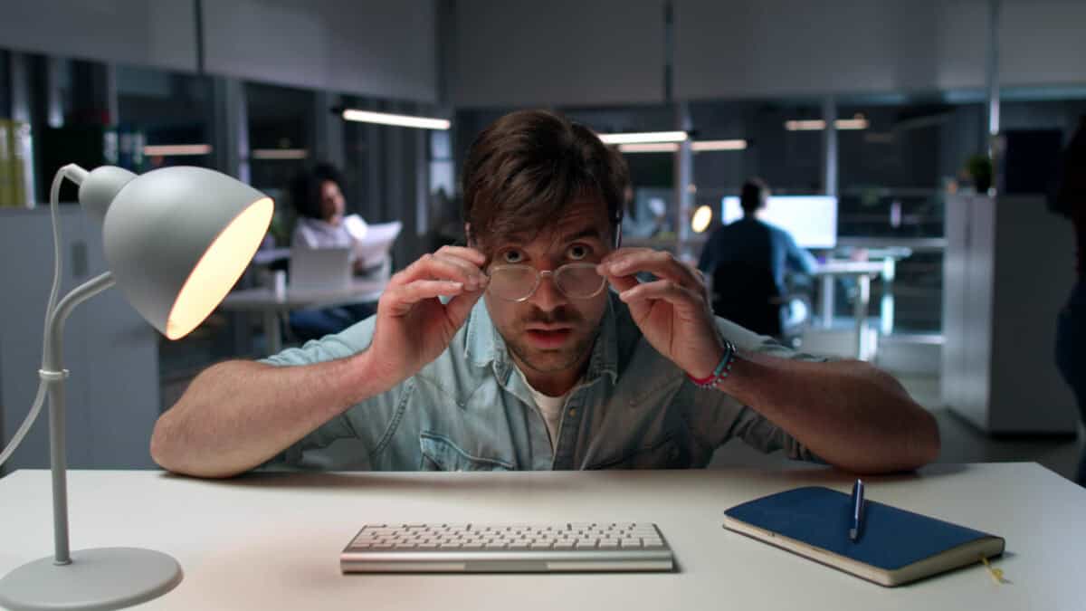 Shocked office worker staring at computer screen with colleagues working in the background.