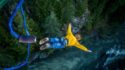 A young person wearing a yellow shirt and jeans dives towards a river below on a bungee cord.