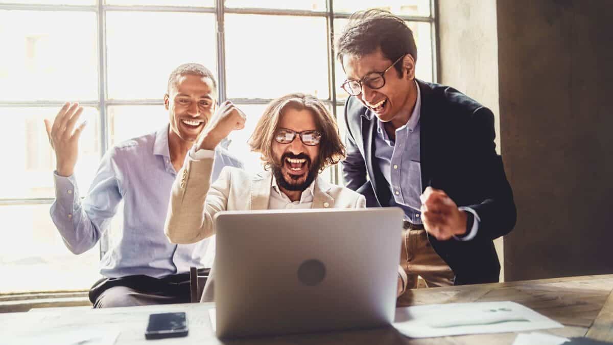 Three happy office workers cheer as they read about good financial news on a laptop.