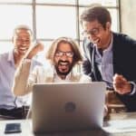 Three happy office workers cheer as they read about good financial news on a laptop.