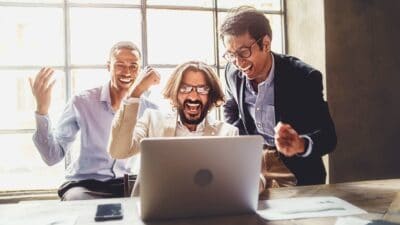 Three happy office workers cheer as they read about good financial news on a laptop.