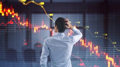 Man with a hand on his head looks at a red stock market chart showing a falling share price.