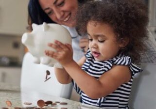 a small girl empties a piggy bank of coins onto a table while her mother looks on in the background.